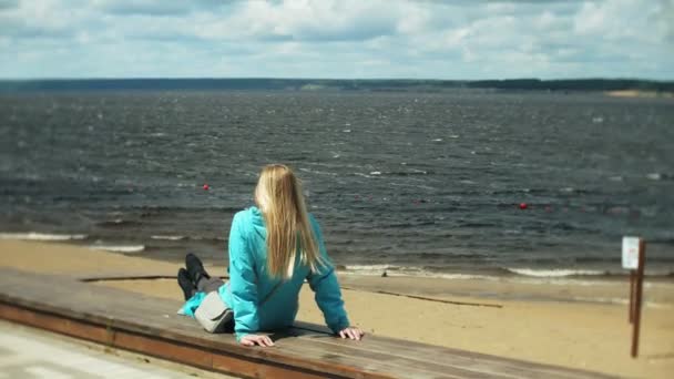 Jeune jolie femme fraîche assise sur une jetée en bois près de la mer et regardant la ville. Attrayant hipster girl avec sac à dos profiter de ses vacances. .Concept de mode de vie actif . — Video