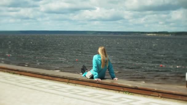 Junge frische hübsche Frau sitzt auf einem hölzernen Steg in der Nähe des Meeres und blickt auf die Stadt. attraktive Hipster-Mädchen mit Rucksack genießen ihren Urlaub. .active Lifestylekonzept. — Stockvideo