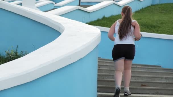 Jonge vet meisje rennen trappen, vergieten gewicht, luisteren naar muziek in hoofdtelefoons concept van gezonde levensstijl super slow motion — Stockvideo