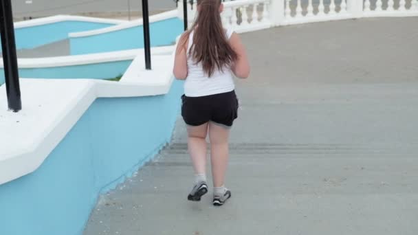 Niña gorda corriendo escaleras abajo, perdiendo peso, escuchando música en los auriculares concepto de estilo de vida saludable super cámara lenta — Vídeos de Stock