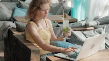 Genç iş kadını masada Mojito kokteyl oturma ve çekici notlar not defterinde içme. Bir dizüstü bilgisayar, akıllı telefon olacak. Öğretim öğrencileri online. Blogger.