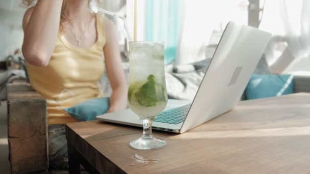 Ung affärskvinna dricka mojito cocktail sittande vid bord och anteckningar i anteckningsboken. Vidare är en laptop, smartphone. Lära elever online. Blogger. — Stockvideo