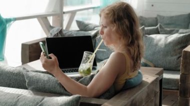 Genç iş kadını masada Mojito kokteyl oturma ve çekici notlar not defterinde içme. Bir dizüstü bilgisayar, akıllı telefon olacak. Öğretim öğrencileri online. Blogger.