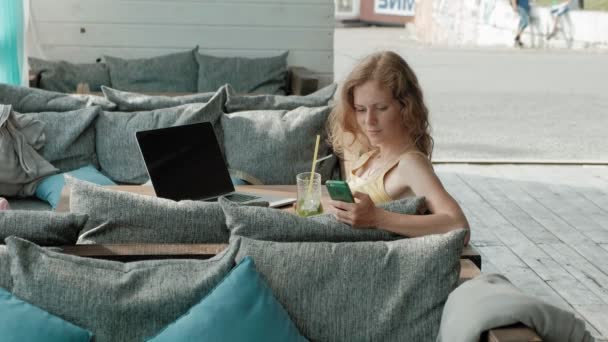 Νεαρή γυναίκα πίνοντας mojito cocktail κάθεται στο τραπέζι και παίρνει σημειώσεις στο Σημειωματάριο. Είναι ένα φορητό υπολογιστή, smartphone. Διδασκαλίας στους μαθητές σε απευθείας σύνδεση. Blogger. — Αρχείο Βίντεο