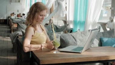 Genç iş kadını masada Mojito kokteyl oturma ve çekici notlar not defterinde içme. Bir dizüstü bilgisayar, akıllı telefon olacak. Öğretim öğrencileri online. Blogger.