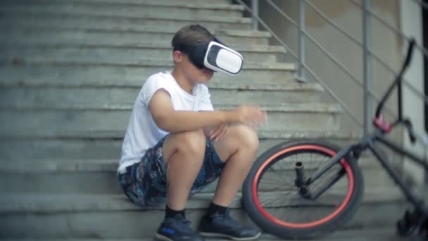 O menino está sentado na escada em óculos virtuais, ao lado da bicicleta BMX — Vídeo de Stock