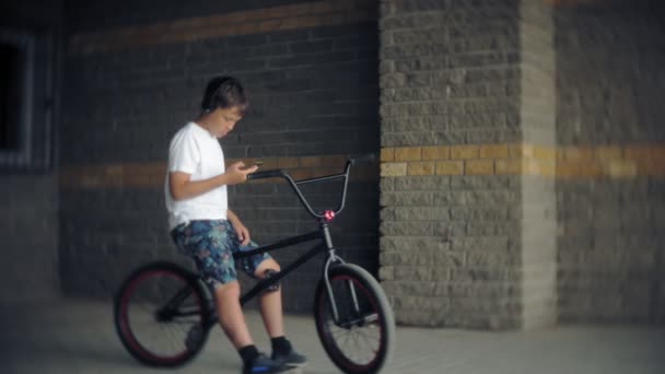 Der Junge sitzt auf einem BMX-Fahrrad und hört Musik von einem Smartphone — Stockvideo