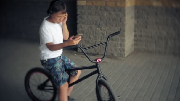 Der Junge sitzt auf einem BMX-Fahrrad und hört Musik von einem Smartphone — Stockvideo