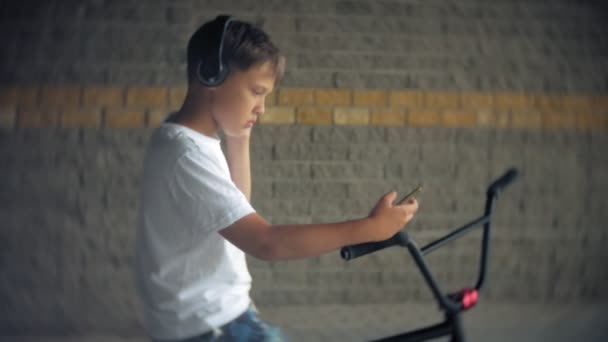 Çocuk bir Bmx bisiklet üzerinde oturur ve bir smartphone--dan müzik dinliyor — Stok video