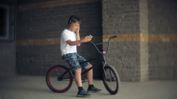 The boy sits on a BMX bike and listens to music from a smartphone — Stock Video