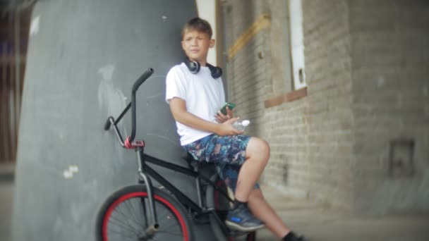 Der Junge sitzt auf einem BMX-Fahrrad und hört Musik von einem Smartphone — Stockvideo