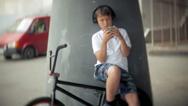 The boy sits on a BMX bike and listens to music from a smartphone — Stock Video