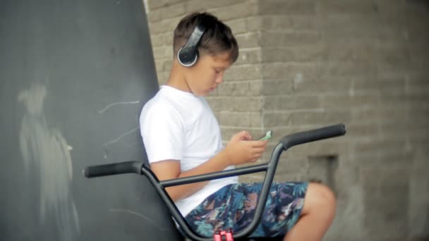 El niño se sienta en una bicicleta BMX y escucha música desde un teléfono inteligente — Vídeo de stock