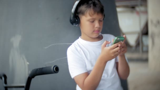 El niño se sienta en una bicicleta BMX y escucha música desde un teléfono inteligente — Vídeo de stock
