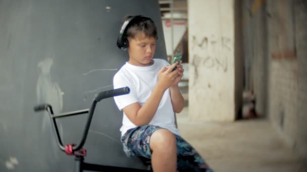 O menino se senta em uma bicicleta BMX e ouve música de um smartphone — Vídeo de Stock