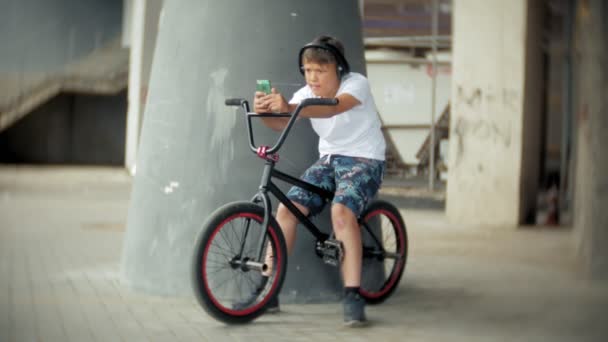Der Junge sitzt auf einem BMX-Fahrrad und hört Musik von einem Smartphone — Stockvideo