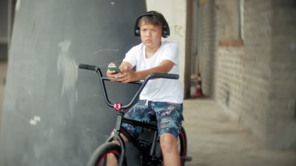 El niño se sienta en una bicicleta BMX y escucha música desde un teléfono inteligente — Vídeos de Stock