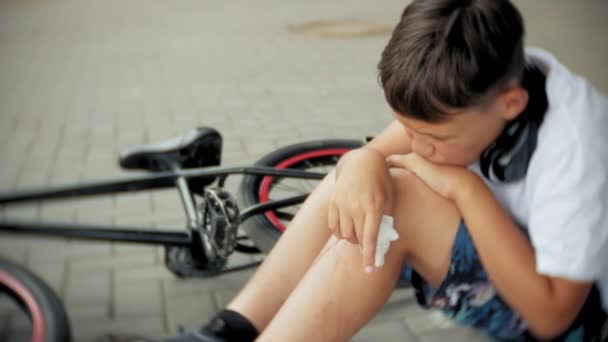 Egy fiatal fiú ül a parkban egy kerékpár esés után, nyugtatja a fájdalom a térde, egy veszélyes biciklizni — Stock videók
