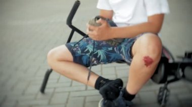 Genç bir çocuk bir parkta bir bisikletten düştükten sonra oturur, onun diz ağrısı yatıştırır, tehlikeli Bisiklete binmek.