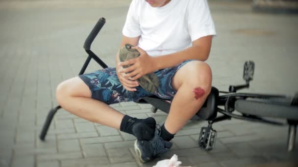 Un jeune garçon est assis dans un parc après être tombé d'un vélo, calme la douleur dans son genou, est une balade dangereuse à vélo — Video