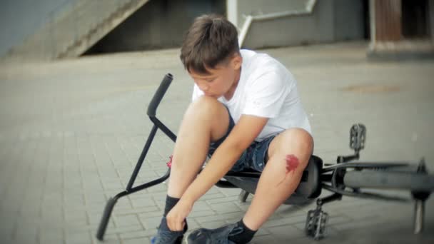 Een jonge jongen in een park zit na het vallen van een fiets, kalmeert de pijn in zijn knie, is een gevaarlijke fietstocht — Stockvideo