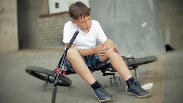 Een jonge jongen in een park zit na het vallen van een fiets, kalmeert de pijn in zijn knie, is een gevaarlijke fietstocht — Stockvideo