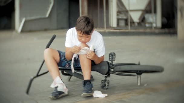 Een jonge jongen in een park zit na het vallen van een fiets, kalmeert de pijn in zijn knie, is een gevaarlijke fietstocht — Stockvideo