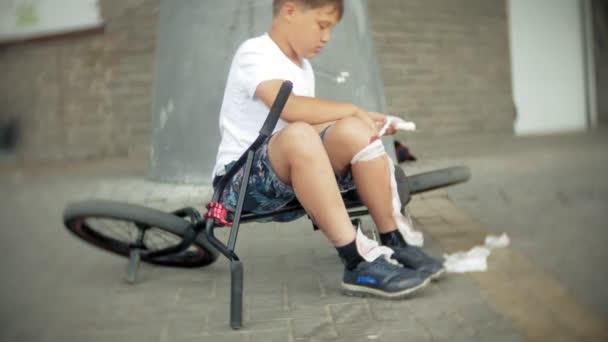 Un niño se sienta en un parque después de caer de una bicicleta, calma el dolor en la rodilla, es un peligroso paseo en bicicleta — Vídeos de Stock