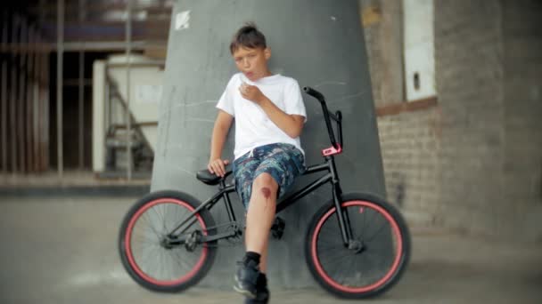 El chico se sienta en una bicicleta BMX y come con apetito de perrito caliente — Vídeos de Stock