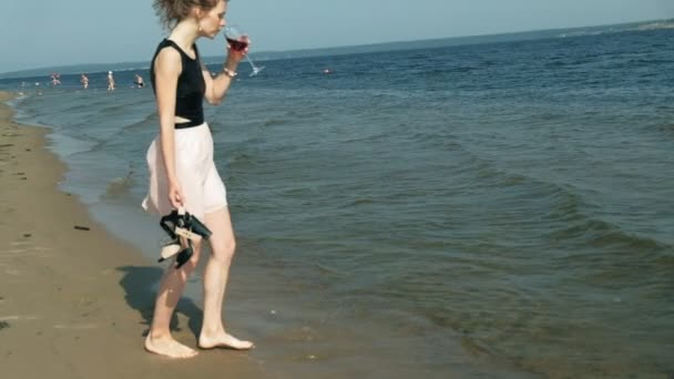 Молодая блондинка в красивом платье наслаждается бокалом розового вина на пляже у моря, супер медленное движение — стоковое видео