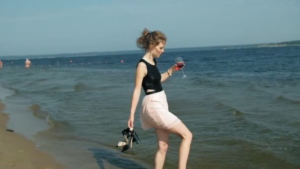 Jovem loira em um lindo vestido desfrutando de um copo de vinho rosa na praia perto do mar, super câmera lenta — Vídeo de Stock