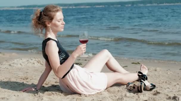 Jeune femme blonde dans une belle robe dégustant un verre de vin rose sur la plage près de la mer, super slow motion — Video