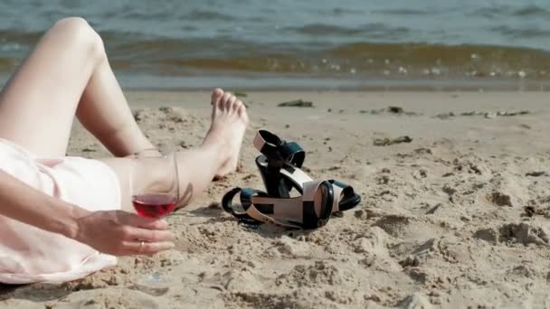 Junge blonde Frau in einem schönen Kleid genießt ein Glas rosa Wein am Strand in der Nähe des Meeres, Super-Zeitlupe — Stockvideo