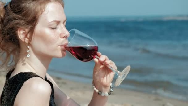 Ung blond kvinna i en vacker klänning som njuter av ett glas rosa vin på stranden nära havet, super slow motion — Stockvideo