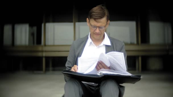 사업가 도시에 있는 계단에 앉아 있다. 그는 양복과 서류 가방을 착용. 그는 문서를 통해 보이는 — 비디오