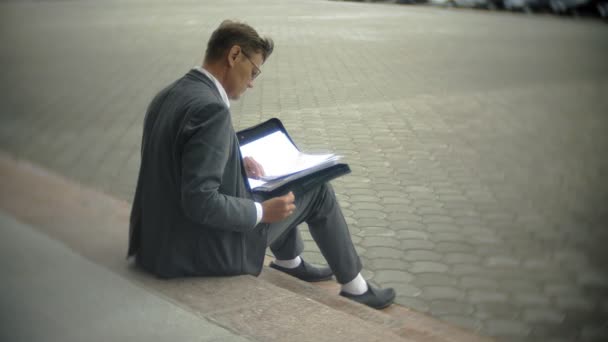 El hombre de negocios está sentado en las escaleras de la ciudad. Lleva traje y maletín. Revisa los documentos. — Vídeos de Stock