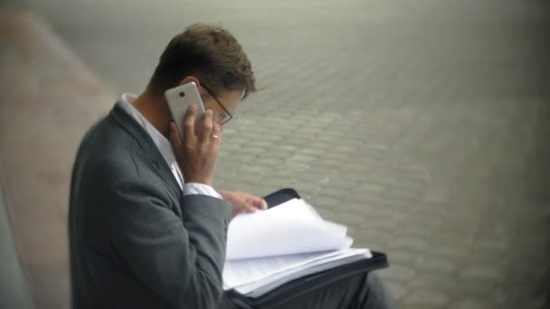 El hombre de negocios está sentado en las escaleras de la ciudad. Lleva traje y maletín. Él mira a través de documentos y conversaciones en el teléfono inteligente — Vídeo de stock