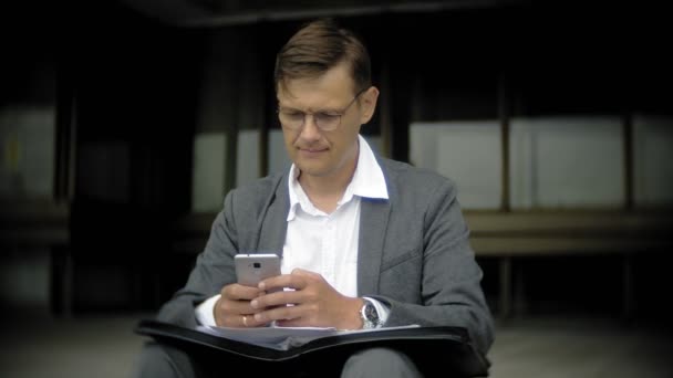 El hombre de negocios está sentado en las escaleras de la ciudad. Lleva traje y maletín. Él mira a través de documentos y conversaciones en el teléfono inteligente — Vídeo de stock