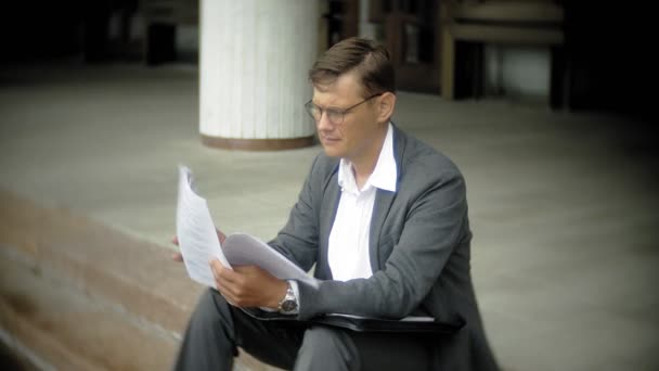 El hombre de negocios está sentado en las escaleras de la ciudad. Lleva traje y maletín. Él mira a través de documentos y conversaciones en el teléfono inteligente — Vídeo de stock