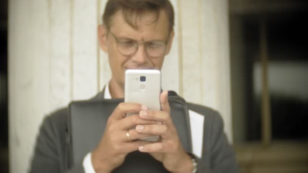 Un homme d'affaires est assis dans les escaliers de la ville. Il porte un costume et une mallette. Il regarde à travers les documents et parle sur le smartphone — Video