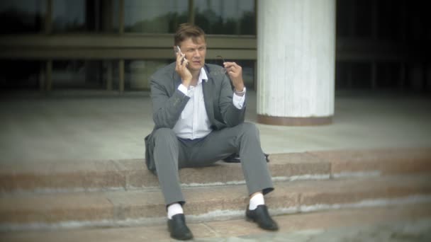 O empresário está sentado nas escadas da cidade. Ele usa um terno e uma pasta e fala no smartphone — Vídeo de Stock