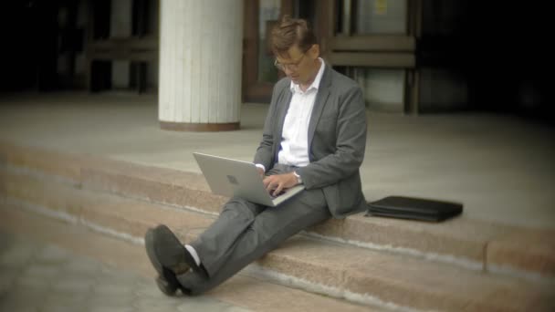 Businessman is sitting on the stairs in the city. He wears a suit and briefcase. He works on a laptop — Stock Video