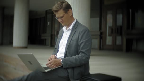 Un homme d'affaires est assis dans les escaliers de la ville. Il porte un costume et une mallette. Il travaille sur un ordinateur portable. — Video