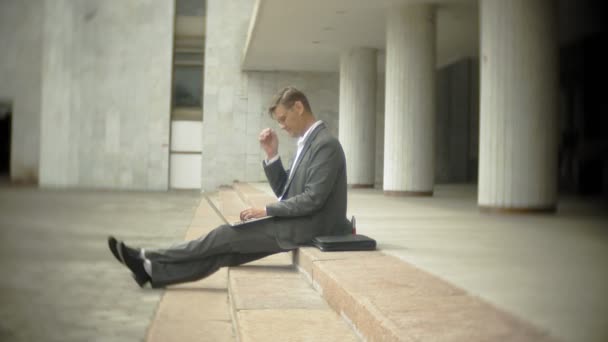 El hombre de negocios está sentado en las escaleras de la ciudad. Lleva traje y maletín. Trabaja en un portátil. — Vídeos de Stock