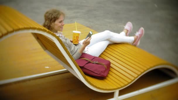 Młoda kobieta z milkshake i telefon komórkowy na zewnątrz na ławce creative komfortowe — Wideo stockowe