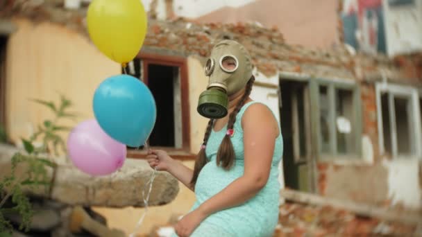 A little girl in a gas mask walks through the ruined buildings with balloons in her hand — Stock Video