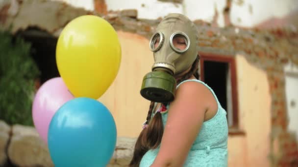 En liten flicka i en gasmask promenader genom de förstörda byggnaderna med ballonger i handen — Stockvideo
