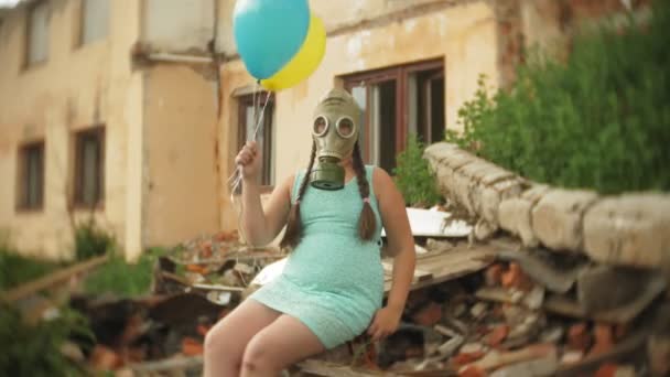 A little girl in a gas mask walks through the ruined buildings with balloons in her hand — Stock Video