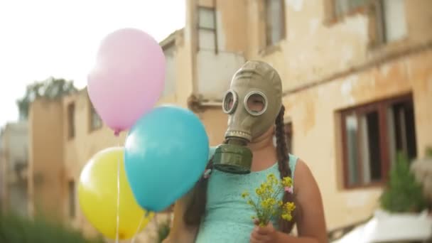 Маленька дівчинка в газовій масці проходить через зруйновані будівлі з повітряними кулями в руці — стокове відео
