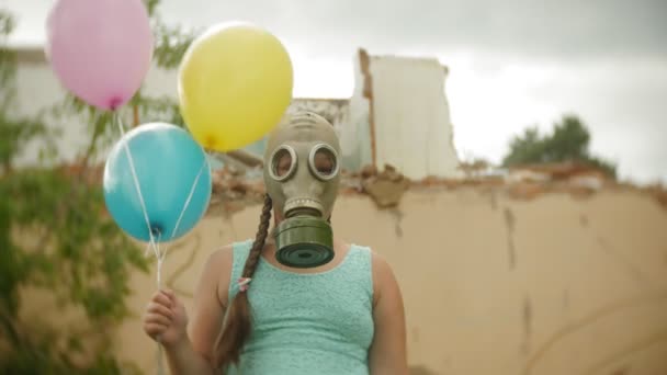 Маленька дівчинка в газовій масці проходить через зруйновані будівлі з повітряними кулями в руці — стокове відео